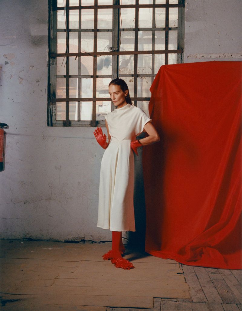 Fashion shot of adult woman in cream dress with red gloves and footwear standing in front of window and red fabric backdrop.
