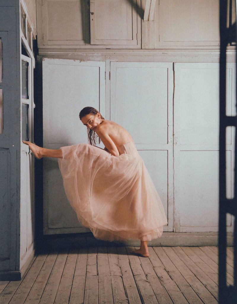 Female dancer in pale pink tulle dress strikes an elegant pose against a vintage corner with gray-painted paneled walls and wood floor.