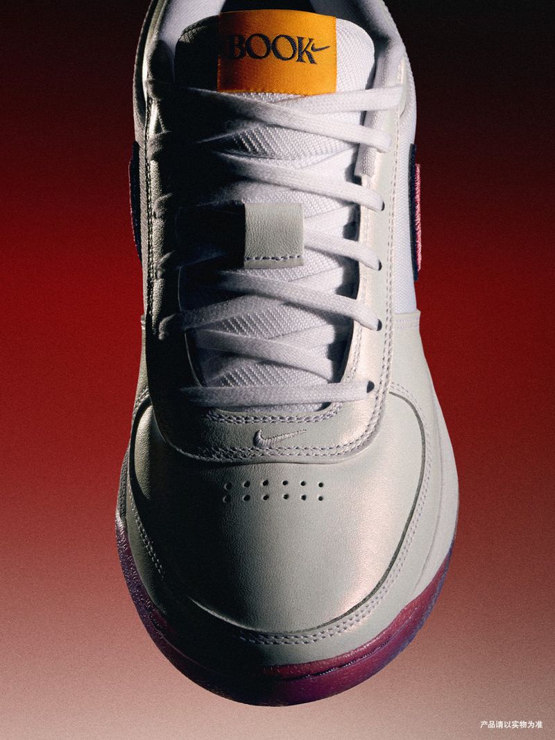 Close-up product shot of a white leather Nike sneaker with white laces, an orange 'BOOK' tag, and a pink sole against a red gradient background.