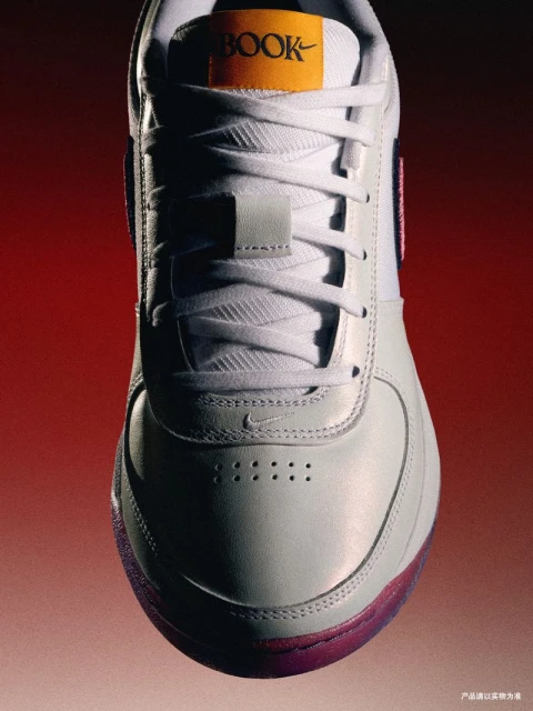 Close-up product shot of a white leather Nike sneaker with white laces, an orange 'BOOK' tag, and a pink sole against a red gradient background.