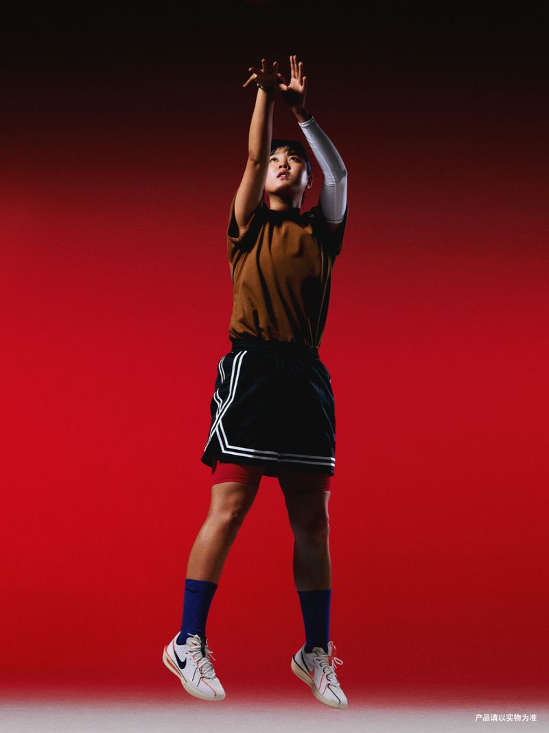 Female basketball player in mid-air, poised to shoot, against a vibrant red background. She's wearing basketball attire and sneakers.