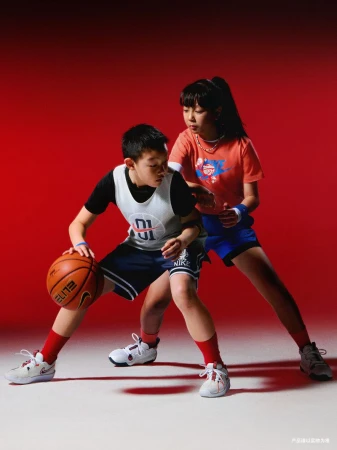 A young boy dribbles a basketball, defended by a slightly older girl on a white surface with a red backdrop. Both are in athletic gear.