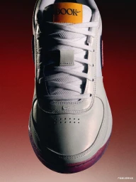 Close-up product shot of a white and grey Nike sneaker with an orange tongue label against a red gradient background.