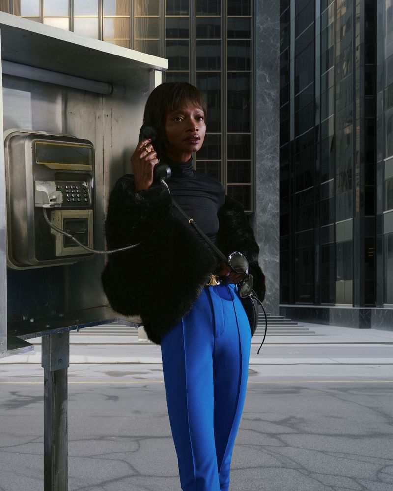 Woman in blue pants and black coat uses payphone in urban setting, surrounded by modern buildings on a grey day.