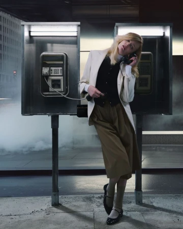 A woman in a white blazer stands next to a phone booth while holding a black telephone receiver in a city with fog and tall buildings.