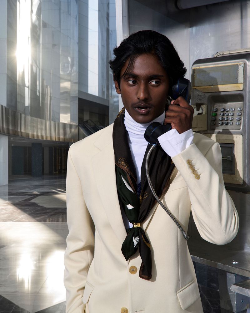 Man in suit using payphone in modern building. He has dark skin and wears a white turtleneck and patterned scarf. 