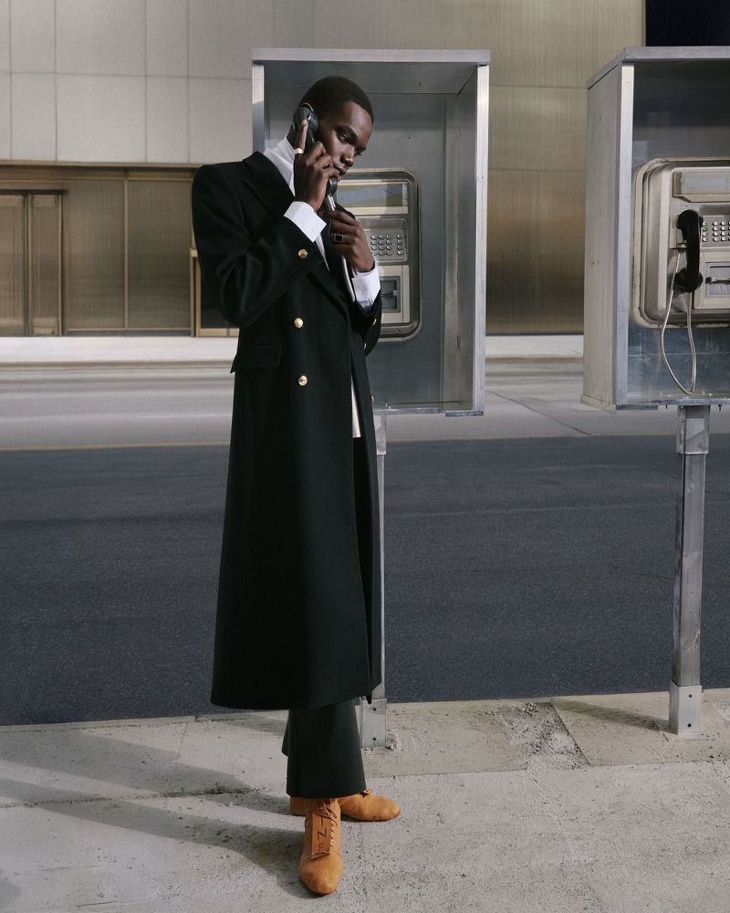 A black man in a long, black overcoat talks on a cell phone next to a row of silver public telephone booths on a city sidewalk.