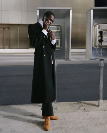 A black man in a long, black overcoat talks on a cell phone next to a row of silver public telephone booths on a city sidewalk.