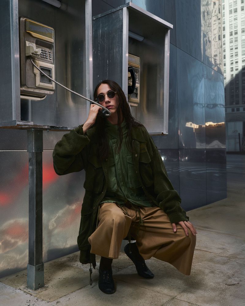 A person using a public payphone in an urban setting, wearing a green jacket, wide brown pants, and black boots, with city buildings in the background.
