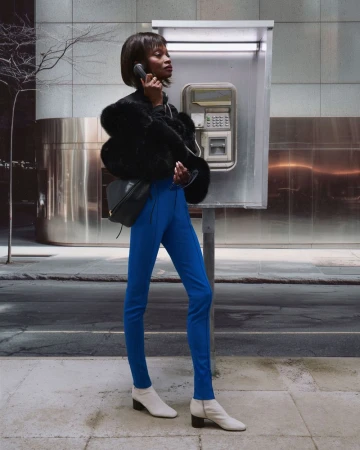 A stylish Black woman poses in cobalt blue pants and white boots while using a payphone on an urban street with a blurred background.