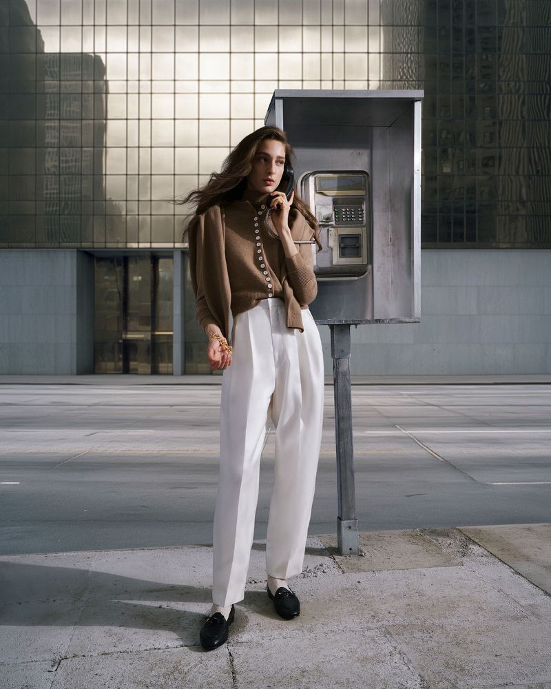 Woman with long brown hair in a brown cardigan and white pants uses a payphone in an urban setting with a modern building behind her.