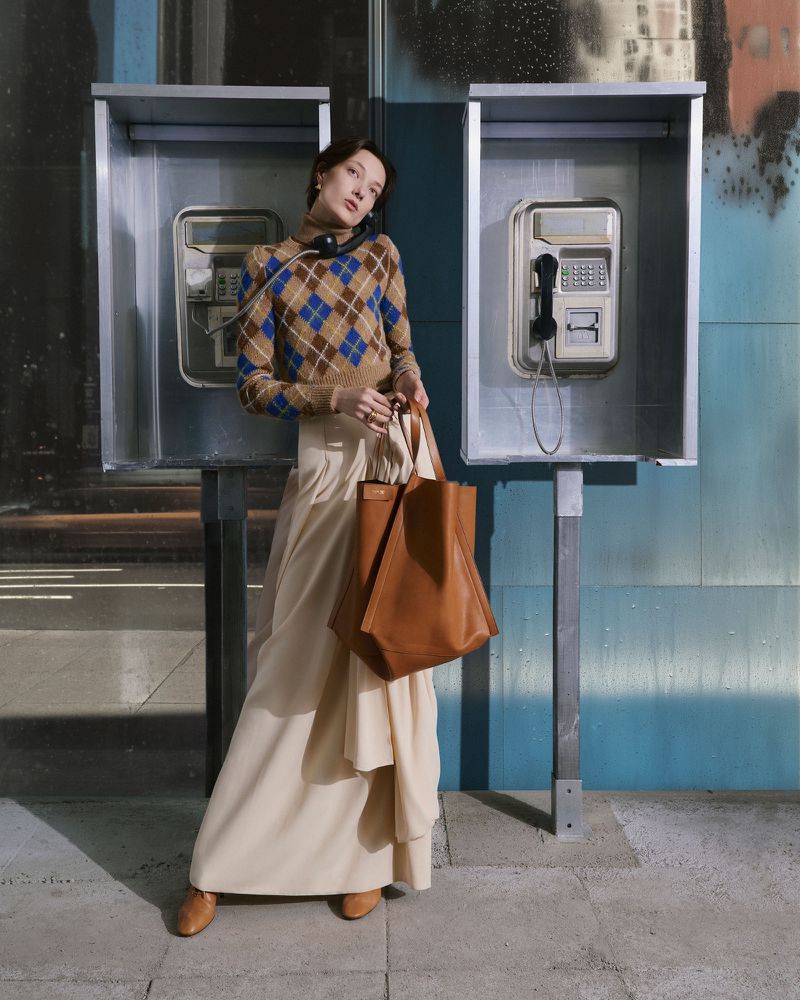 Adult woman poses in front of two telephone booths wearing a diamond-pattern sweater, long skirt, brown shoes, and carrying a brown tote.