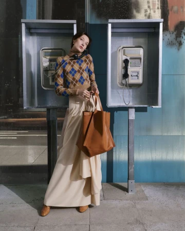 Adult woman poses in front of two telephone booths wearing a diamond-pattern sweater, long skirt, brown shoes, and carrying a brown tote.