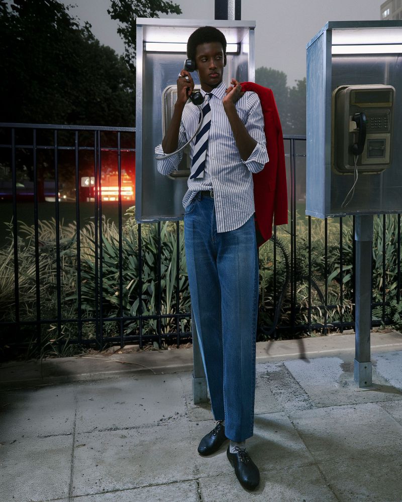 A stylish African-American man stands near a public phone booth at night, wearing a blue shirt, tie, jeans and carrying a red jacket.
