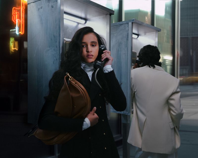 Adult woman in a pea coat uses a vintage telephone in an outdoor phone booth, city street background, and another figure in the distance.