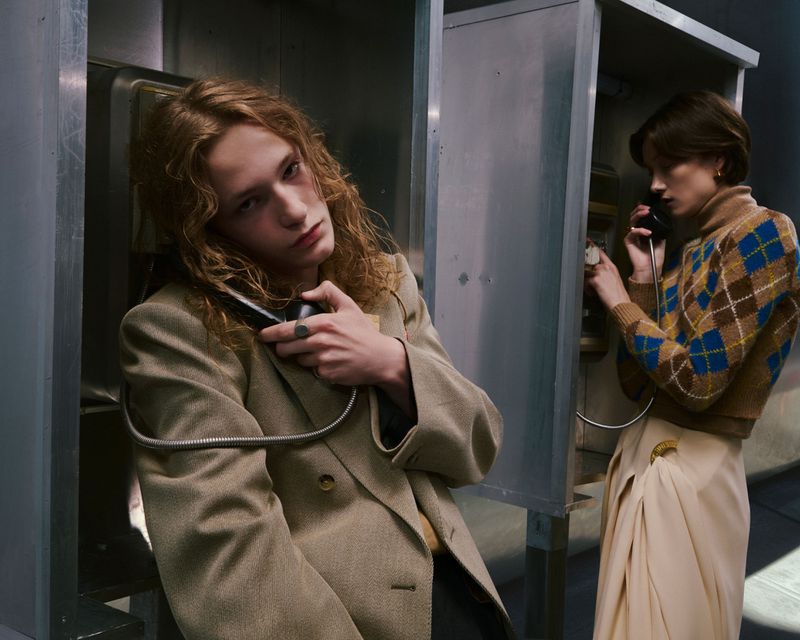 Two models in phone booths: one with long curly hair in a beige blazer, and another with a brown argyle sweater talking on the phone.