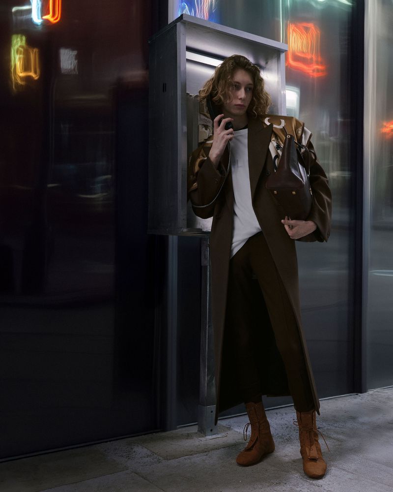A person stands next to a phone booth wearing a brown suit, white shirt, and brown boots against a reflective background with neon signs.