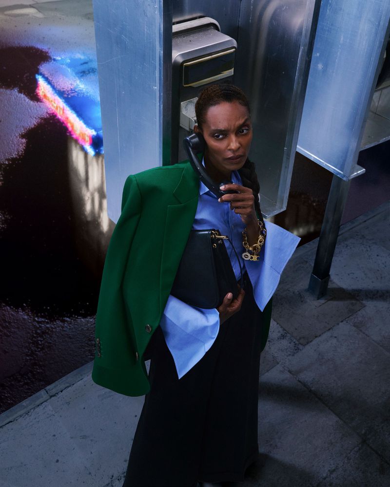 A dark-skinned woman in a green blazer stands in a phone booth, holding a purse and talking on the phone, lit by colorful city lights.