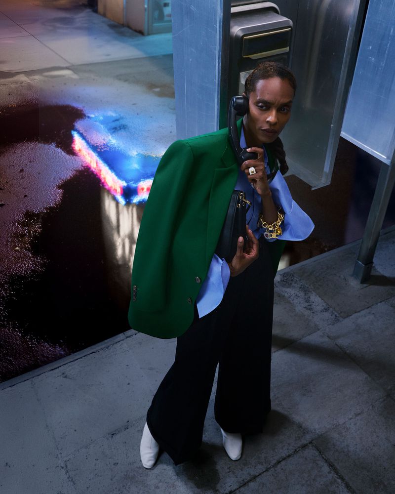 A stylish person in a green blazer, jewelry and black pants, is using a public pay phone on a city street at night. 