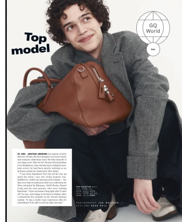 Fashion editorial shot featuring a young adult male in a grey coat holding a brown leather duffel bag, GQ magazine advertisement.