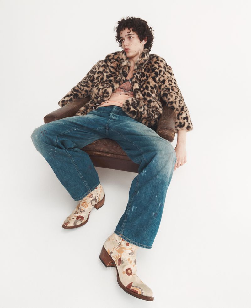 A young adult male sits in a chair wearing a leopard print coat, blue jeans, and floral print boots against a white backdrop.