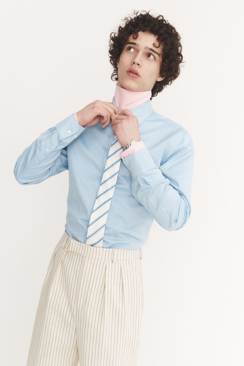 A young adult male model with curly dark hair wears a light blue shirt and striped pants, adjusting his tie in a light studio.
