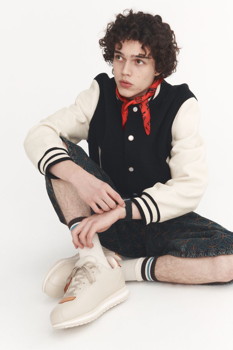 Young adult with curly hair poses while sitting on a white floor in a varsity jacket, red bandana, and sneakers in a studio setting.