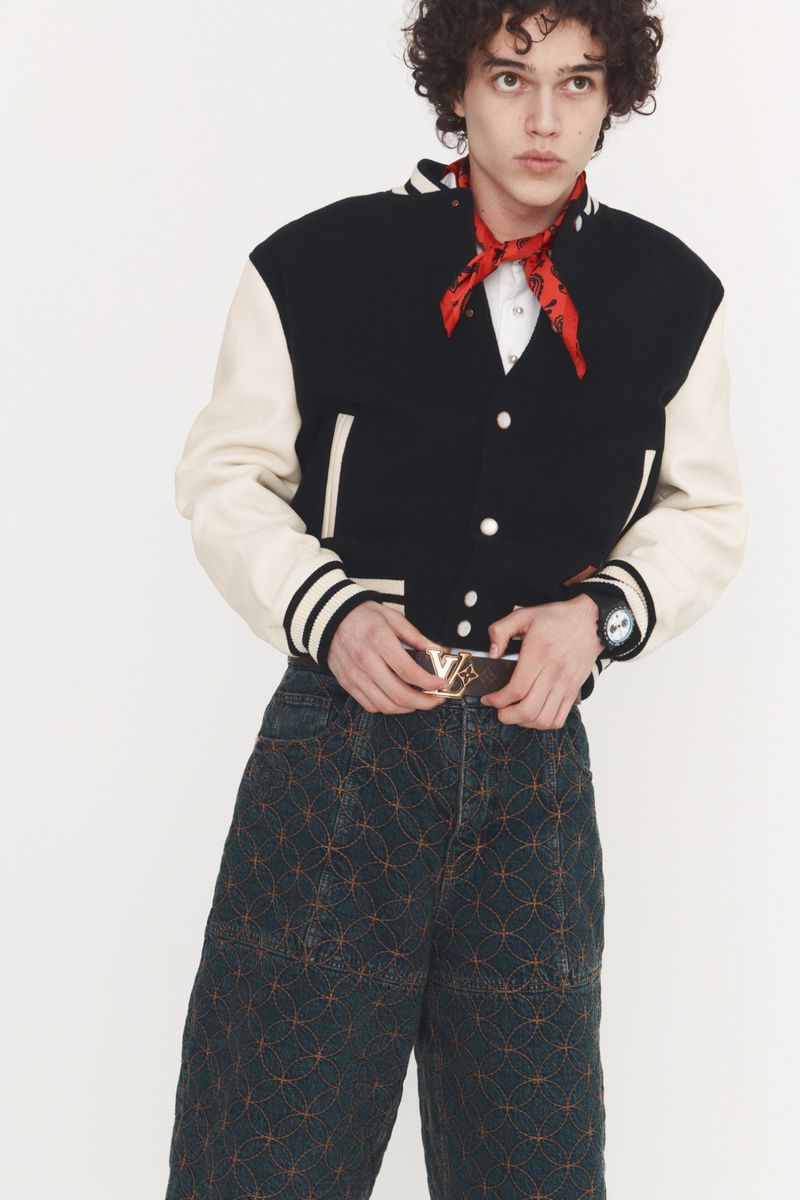 Young man in a varsity jacket and patterned pants poses against a white background in a fashion editorial photograph.