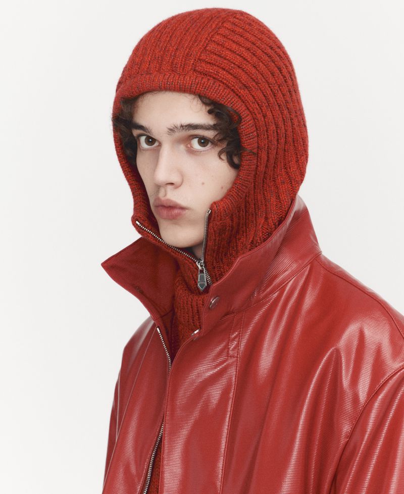 Close-up of a young adult with curly hair, wearing a red hooded balaclava and a matching red vinyl jacket against a white background.