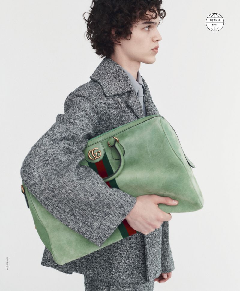 Adult male with curly hair wears a gray tweed suit while holding a large green Gucci duffel bag against a plain white background.