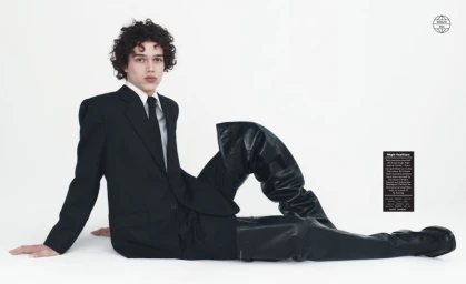 Model Cruz Kahlbetzer in a black suit and oversized black leather thigh-high boots reclining on the floor against a white backdrop.