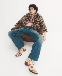 Model Cruz Kahlbetzer seated in an armchair wearing a leopard print coat, denim jeans, and patterned boots on a white backdrop.