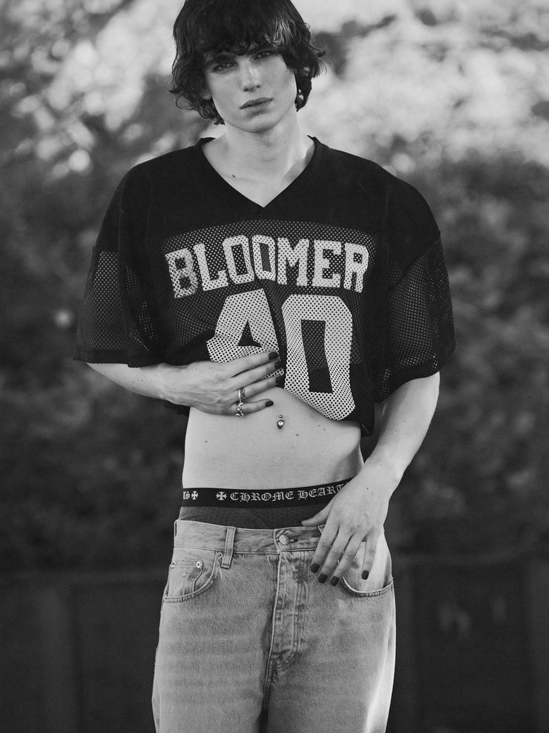 Black-and-white portrait of a slender young man in an open sports jersey, jeans, and visible boxer waistband against a blurred backdrop.