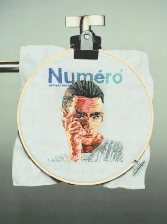 Embroidery art featuring a man's portrait and 'Numéro' logo, displayed on a towel rack, showcasing textile art and typography.