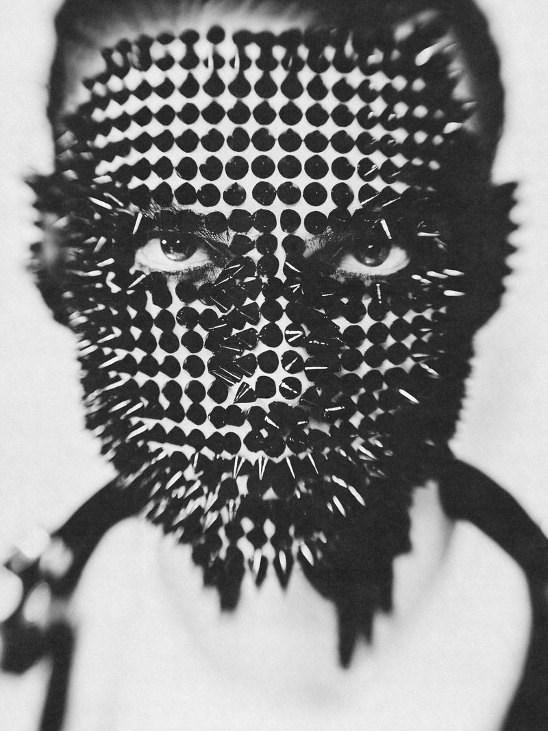 Monochrome portrait of a person wearing a spiked mask, intense gaze, minimalist background, and high-contrast aesthetic. Close-up.