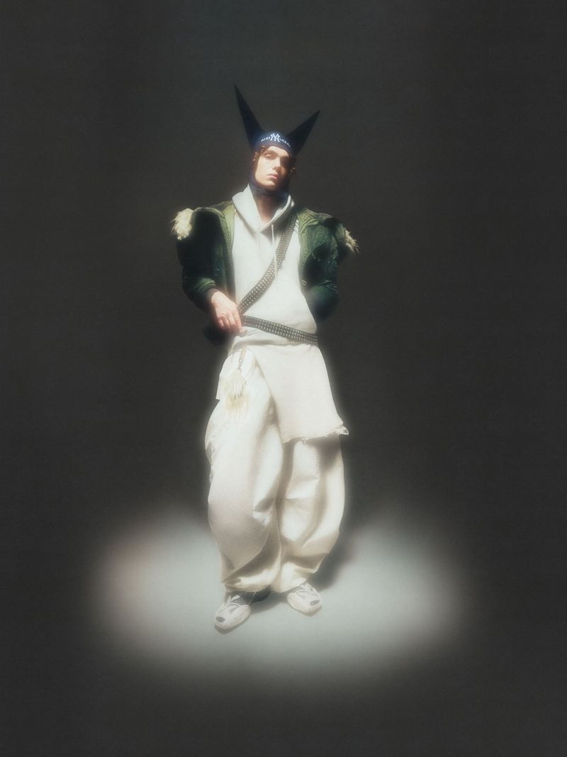 Person in avant-garde fashion with a peaked hat, wearing a hooded top and loose pants, spotlit against a dark grey backdrop. 