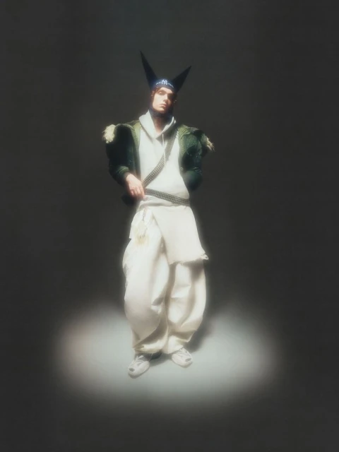 Person in avant-garde fashion with a peaked hat, wearing a hooded top and loose pants, spotlit against a dark grey backdrop. 