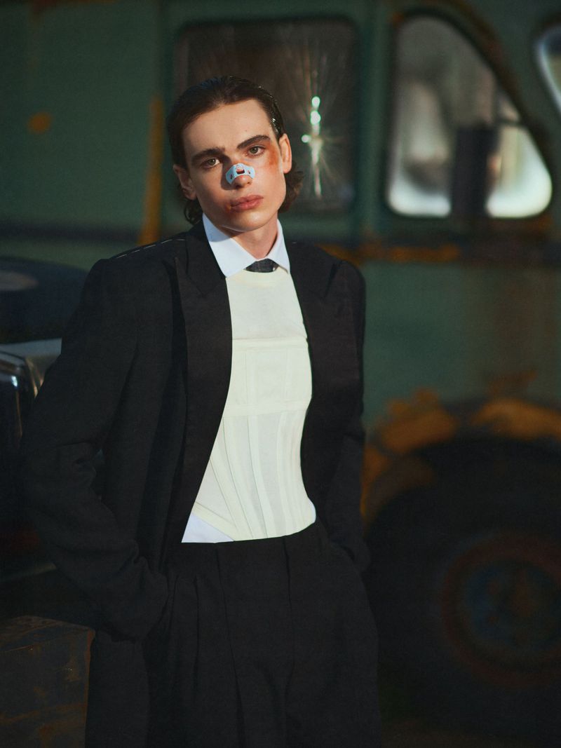 Adult male in formal attire with facial injury stands in front of rusted truck in moody light, posing for fashion editorial photography.