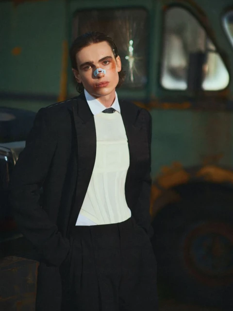 Adult male in formal attire with facial injury stands in front of rusted truck in moody light, posing for fashion editorial photography.
