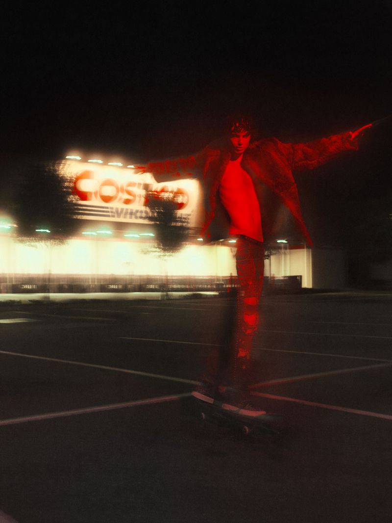 Adult male skateboarding in parking lot near brightly lit commercial building at night; red lighting with motion blur effect.