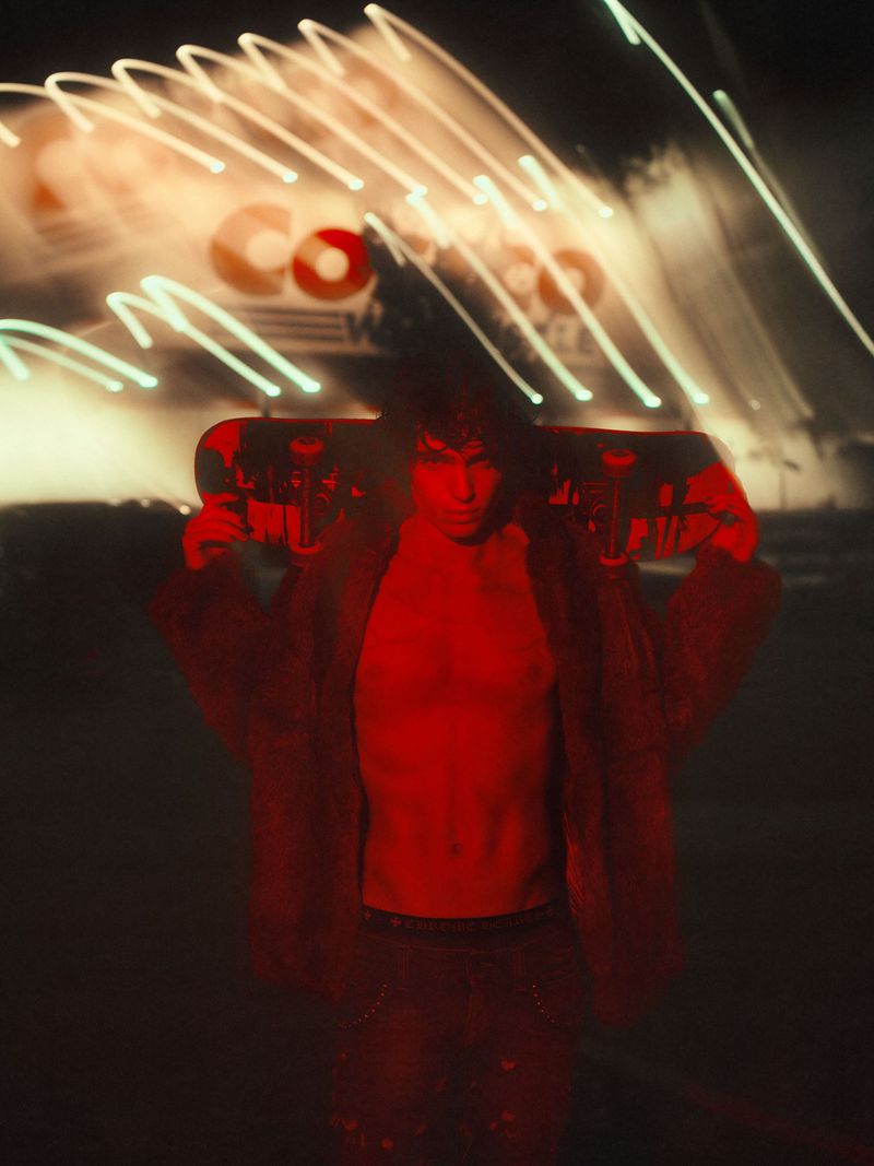 An adult male with dark hair holds a skateboard over his shoulders, shot with long exposure, red lighting, and a dark urban setting.