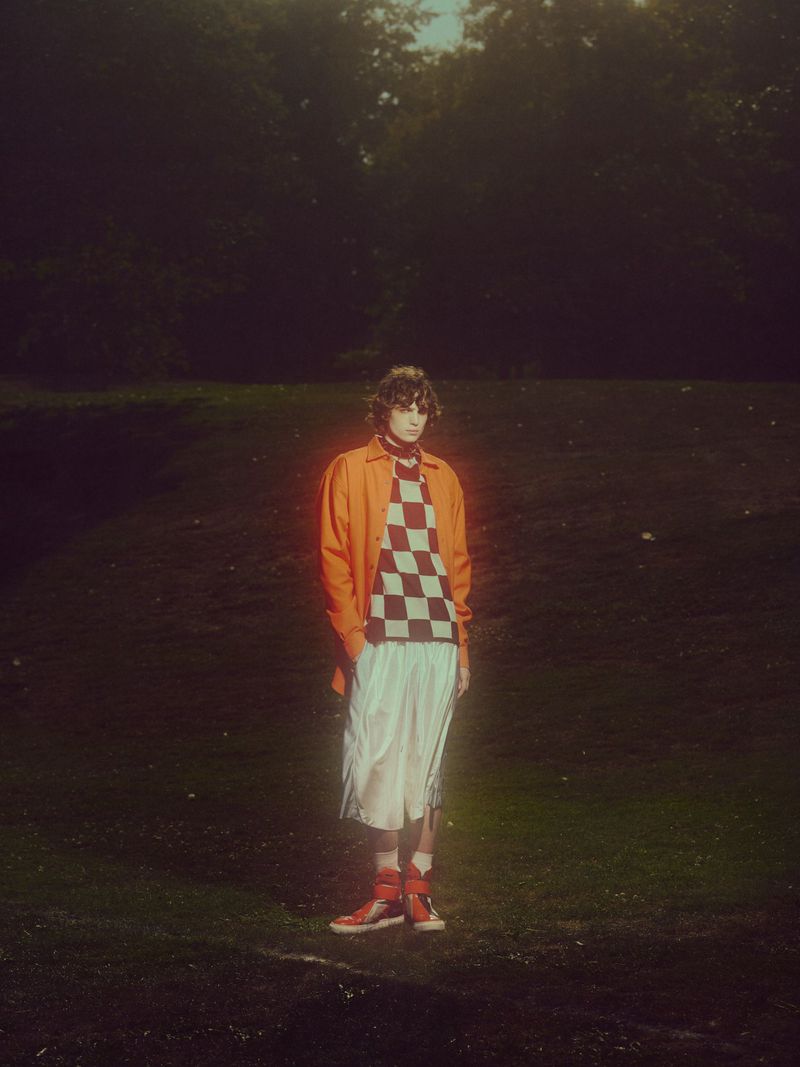 Young adult in an orange jacket and checkered shirt stands in a field with trees in background, captured with natural lighting.