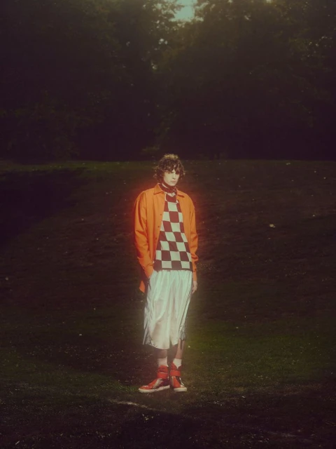 Young adult in an orange jacket and checkered shirt stands in a field with trees in background, captured with natural lighting.