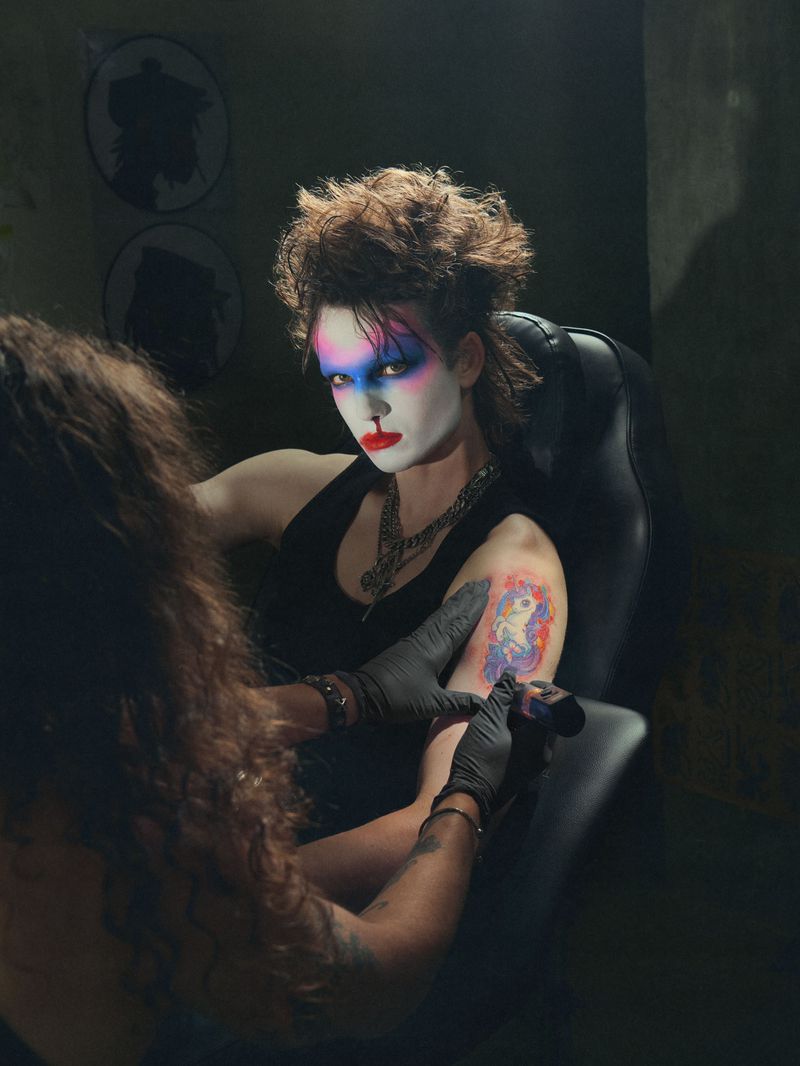 Woman with striking makeup in a chair gets a unicorn tattoo, black gloves on the tattoo artist, dark atmosphere, unique style.