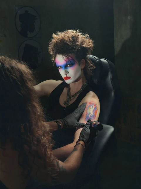 Woman with striking makeup in a chair gets a unicorn tattoo, black gloves on the tattoo artist, dark atmosphere, unique style.