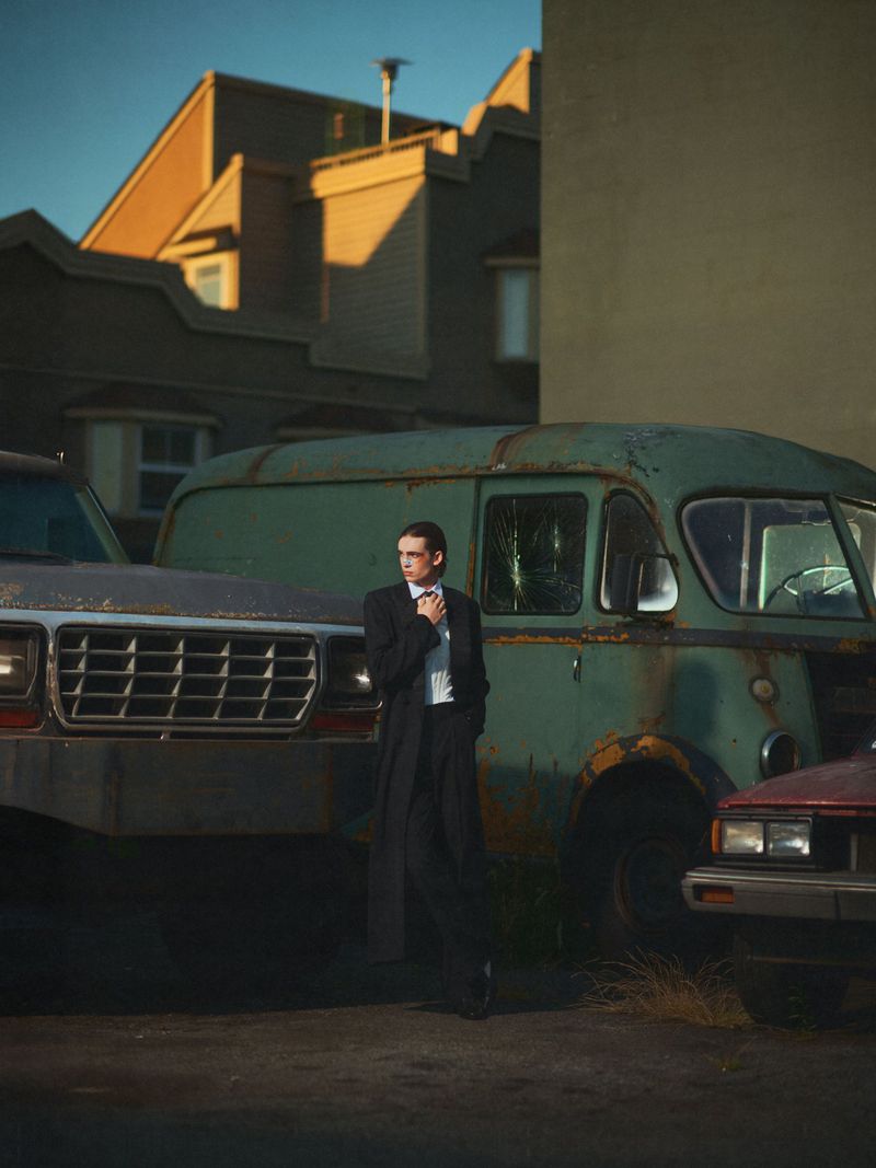 An adult man in a suit stands in a parking lot near several old vehicles, including a rusted van, under a clear sky at golden hour.