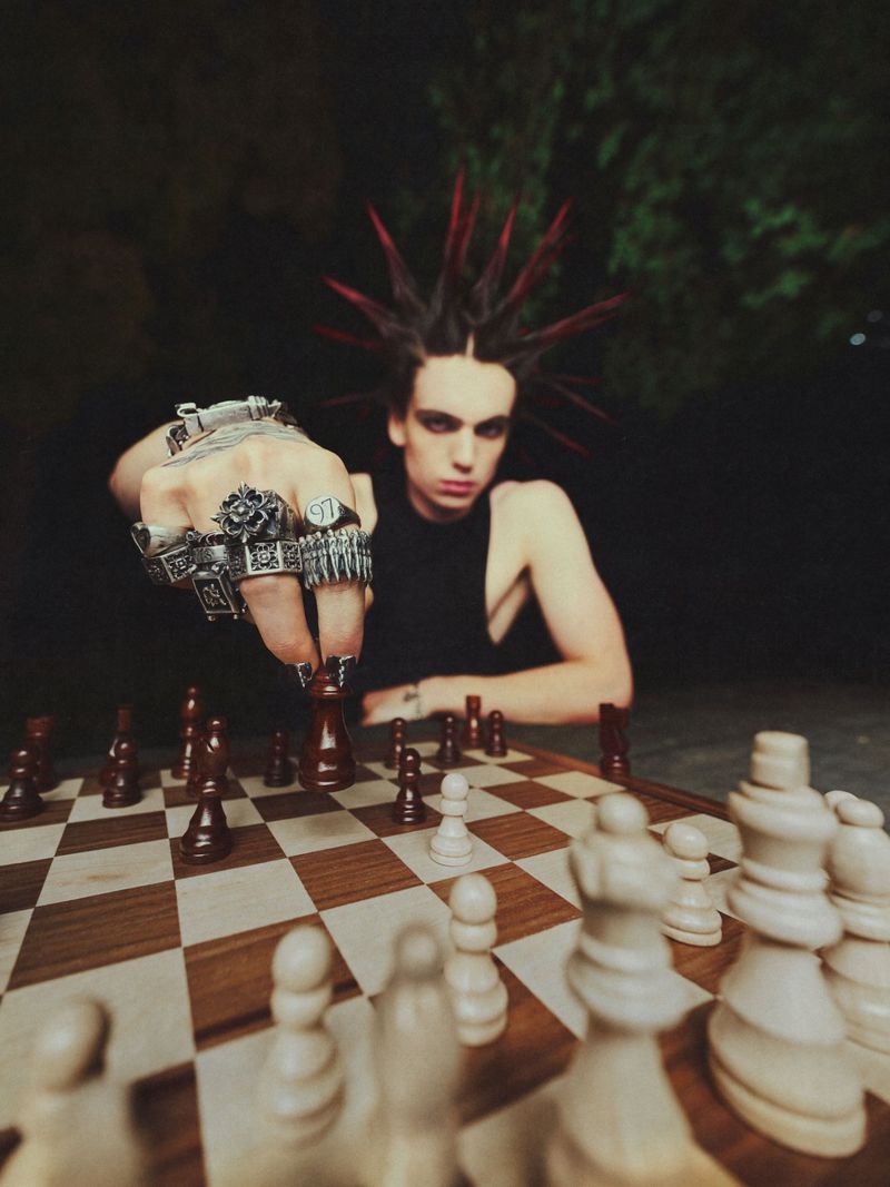 A young adult with a punk hairstyle wearing rings is playing chess, with a dark background and dramatic lighting, creating a stylish portrait.