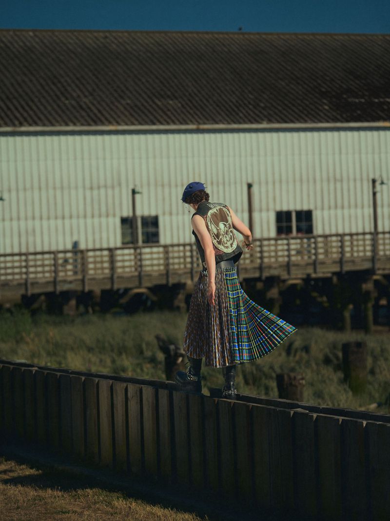 Young adult woman in vest and kilt balances on wooden fence outdoors against building in diffused daylight. Fashion editorial shoot.