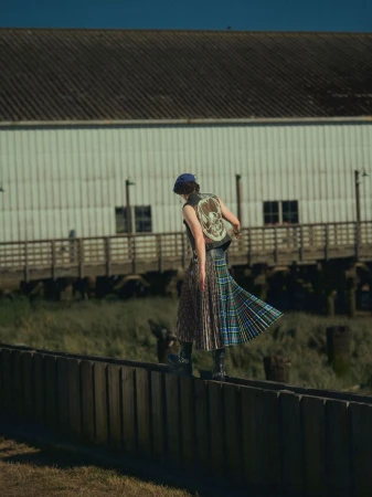 Young adult woman in vest and kilt balances on wooden fence outdoors against building in diffused daylight. Fashion editorial shoot.