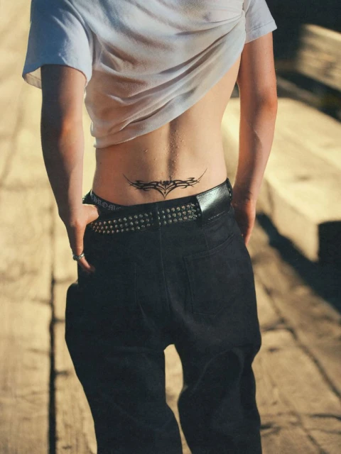 Person in low-rise jeans with studded belt shows a lower back tattoo while standing outdoors in casual clothing.